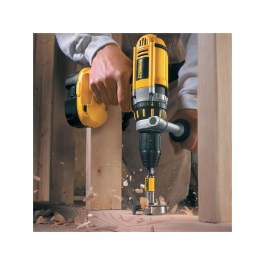 A person using a DEWALT cordless drill with a DEWALT drill bit to drill a hole through a wooden board, with wood shavings flying.