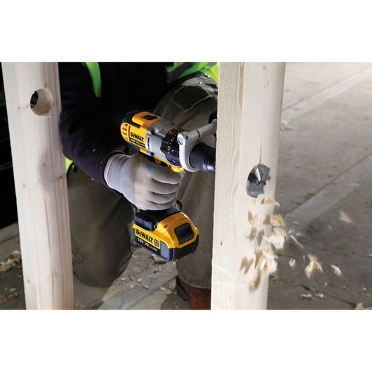 A person using a DEWALT cordless drill to bore a large hole through a wooden beam, with wood chips flying out on a construction site.
