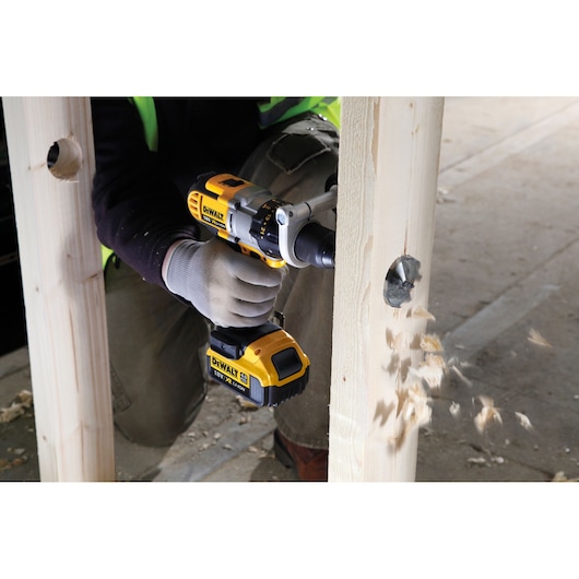 A person using a DEWALT cordless drill to bore a large hole through a wooden beam, with wood chips flying out from the drilled area.