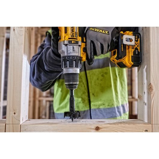 Person uses DEWALT drill to make a hole in wooden frame at construction site, wearing gloves and safety jacket.