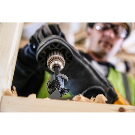 Close-up of a DEWALT DT4582 drill bit in use drilling through wood, held by a gloved hand.