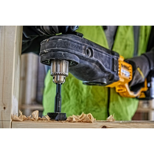 Close-up of DEWALT drill making a hole in wood, wood shavings visible, person in green vest operating tool.