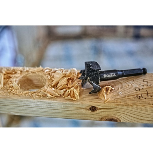 DEWALT drill bit with wood shavings and a drilled hole on a wooden plank in a workshop setting.