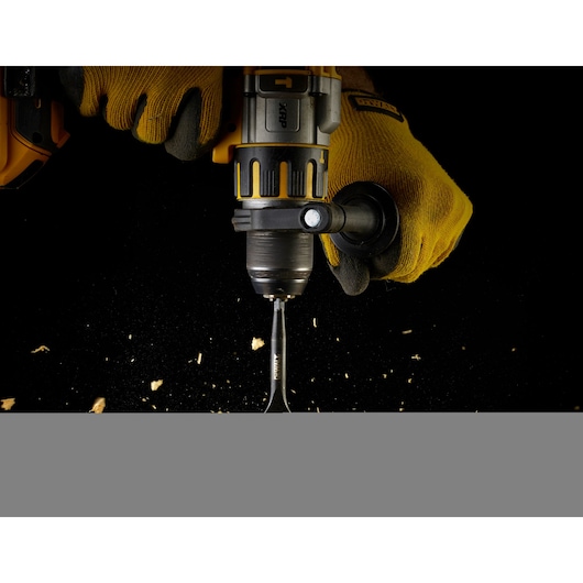 Close-up of a person wearing yellow gloves using a DEWALT power drill with a flat wood drill bit, drilling into a dark surface with wood shavings flying.