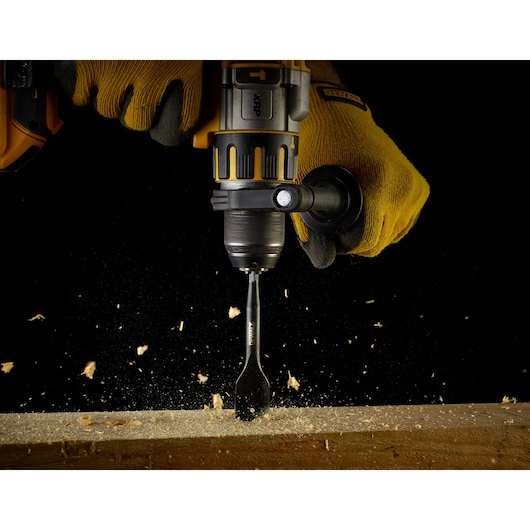 A close-up view of a gloved hand operating a DEWALT drill with a DEWALT flat wood drill bit, drilling into a piece of wood. Wood chips are flying around, indicating active drilling.