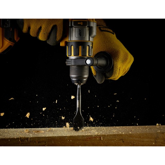Close-up of a DEWALT power drill being used with a flat wood drill bit, operated by a person wearing yellow work gloves. Wood chips and sawdust are flying as the bit drills into a wooden surface.