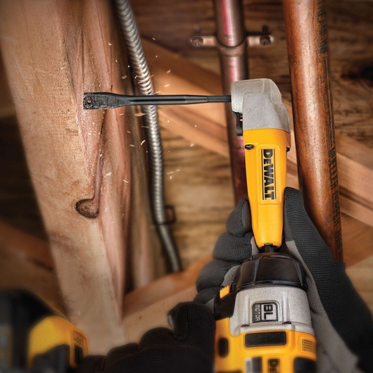 A person wearing black gloves is using a DEWALT power drill with an attachment to chisel or cut into a wooden beam. There are wood chips and dust showing active work. The tool is being used in a tight space near metal pipes.