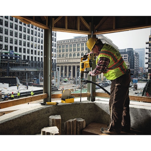 A construction worker using a DEWALT power drill to drill into concrete at a building site in an urban area. The worker is wearing a yellow safety helmet and a high-visibility vest. Other workers and construction activities are visible in the background.