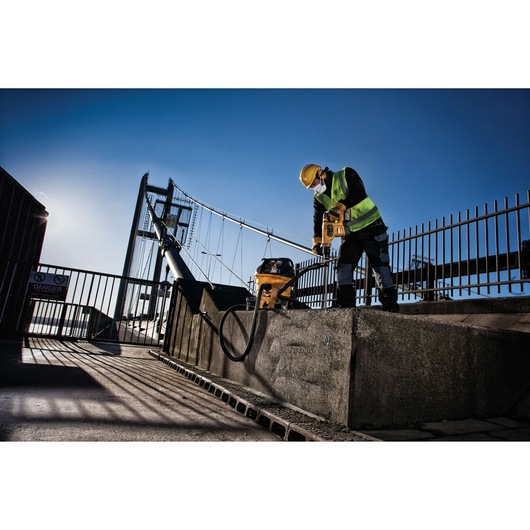 A construction worker wearing a yellow hard hat, high-visibility vest, and face mask uses a DEWALT power tool on a concrete surface near a bridge. The worker is accompanied by a DEWALT vacuum system, with a clear blue sky and structural elements of the bridge visible in the background.