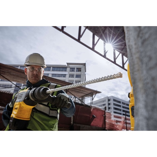 Worker uses DEWALT rotary drill with DT60811 masonry bit at a construction site, with buildings in the background.