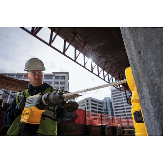 Worker uses a DEWALT drill at a construction site with buildings and safety gear visible in the background.