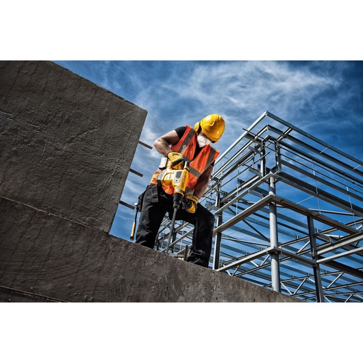 Worker in safety gear uses yellow tool on concrete at construction site with steel framework under blue sky.