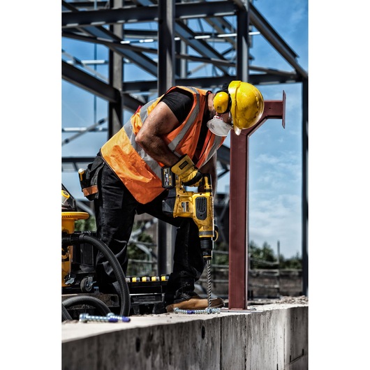 A construction worker wearing an orange safety vest, yellow hard hat, ear protection, and a mask is using a yellow DEWALT power drill to drill into concrete at a building site with exposed steel framework.