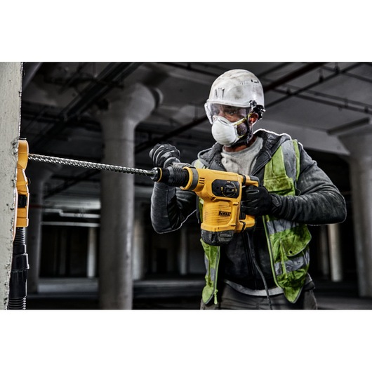 A construction worker wearing a helmet and safety vest uses a yellow DEWALT rotary hammer drill to drill into a concrete pillar in an industrial setting.