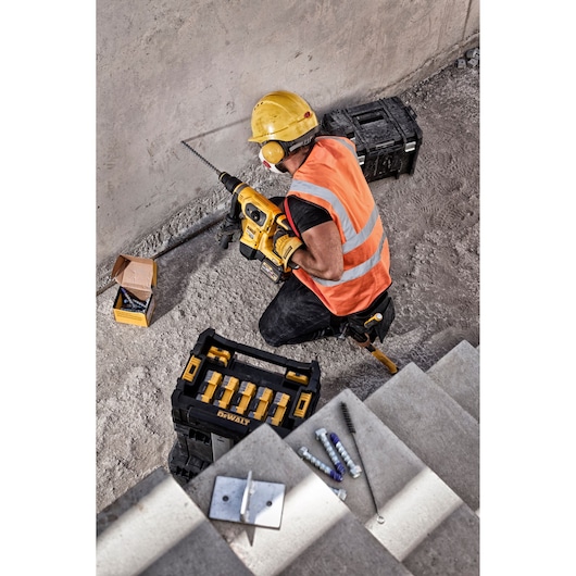 A person wearing a yellow hard hat and orange safety vest is using a DEWALT power tool to drill into a concrete wall at a construction site. Various construction items, including screws, drill bits, a tool box, and a black storage box, are visible around them.