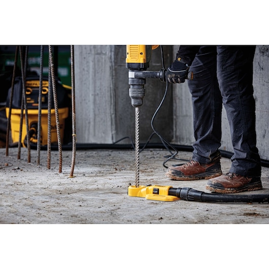 A person using a yellow DEWALT hammer drill to drill into a concrete floor at a construction site. The drill is connected to a dust extraction device. Rebar rods are visible nearby and the person is wearing work boots and gloves.