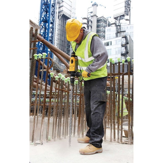 A construction worker wearing a yellow hard hat, a high-visibility vest, gloves, and work boots uses a DEWALT power tool to drill into concrete at a building site surrounded by steel rebar.