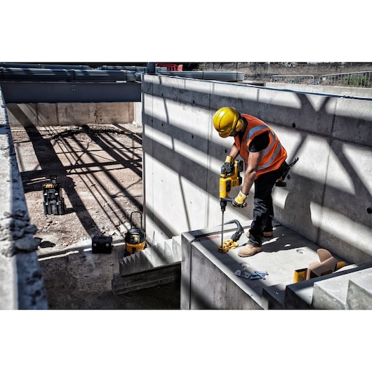 A construction worker wearing a yellow hard hat and orange safety vest uses a DEWALT power tool to drill into a concrete structure at an industrial construction site. Various tools and equipment are visible in the background.
