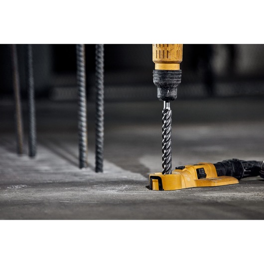 A yellow DEWALT drill with a spiral drill bit is being used to drill into a concrete surface. Metal rods are visible in the background, indicating a construction environment.