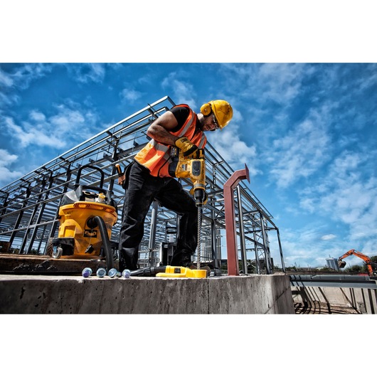 A construction worker wearing a yellow hard hat and orange safety vest uses a DEWALT power drill on a concrete surface at an industrial building site. A yellow DEWALT vacuum is nearby, and metal structural framework is visible in the background under a bright blue sky.