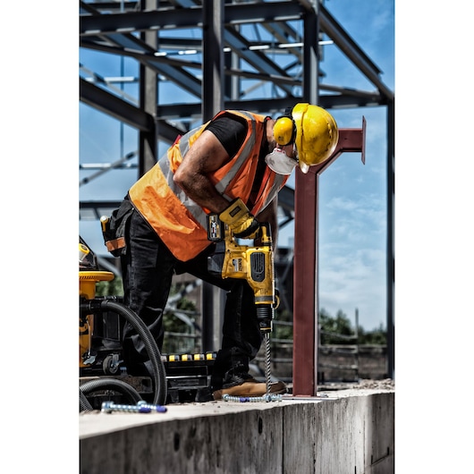 A construction worker wearing a yellow hard hat, orange safety vest, and face mask is using a DEWALT power drill to drill into a concrete surface at an outdoor construction site. Steel framework is visible in the background.