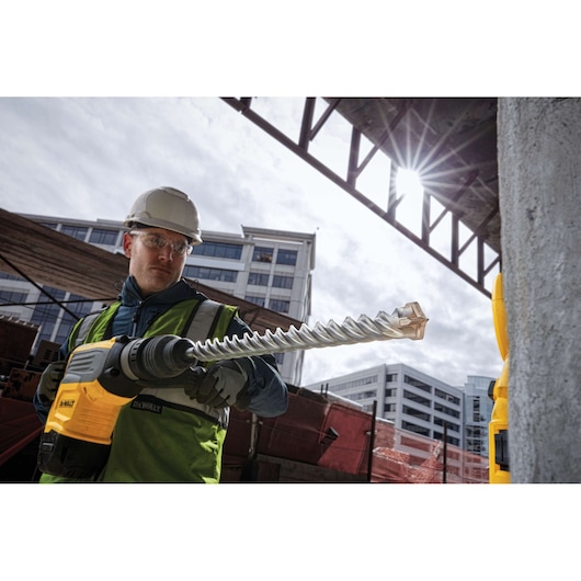 A construction worker wearing a safety helmet and vest uses a DEWALT power tool fitted with a large spiral drill bit on a building site. Modern buildings and construction materials are visible in the background.