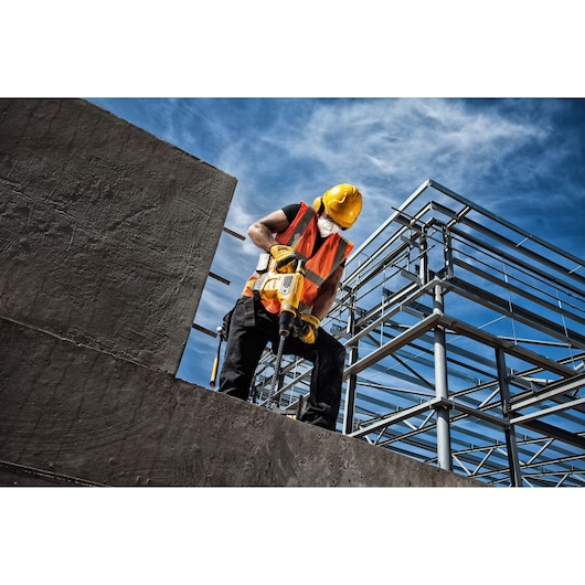 A construction worker wearing a yellow helmet, orange safety vest, and face mask uses a yellow DEWALT power tool to drill into a concrete structure at an industrial or construction site with metal framework in the background.