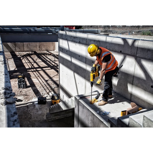 A construction worker wearing a yellow hard hat and an orange safety vest uses a DEWALT power tool to drill into a concrete wall at a construction site. Several DEWALT tool cases and equipment are visible on the ground.