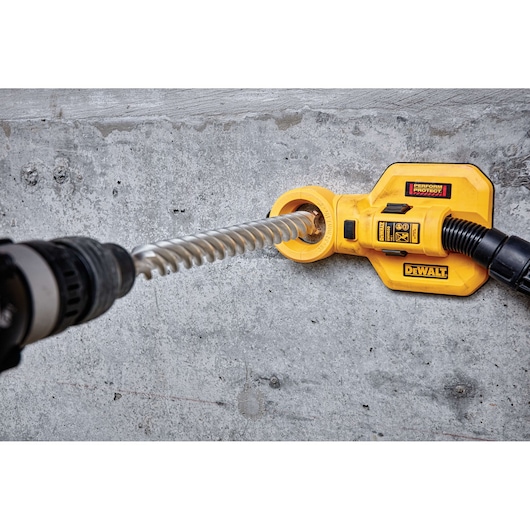 A DEWALT drill attachment is in use on a concrete wall, with a spiral drill bit inserted and dust extraction hose connected.