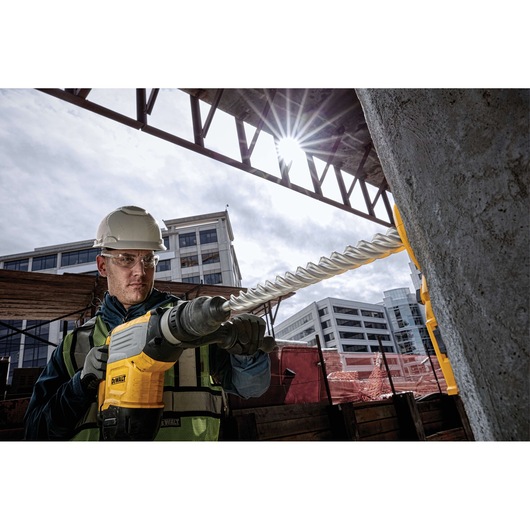 A person wearing a safety helmet and vest operates a DEWALT power tool with a large drill bit, drilling into a concrete pillar at a construction site. Modern buildings can be seen in the background.