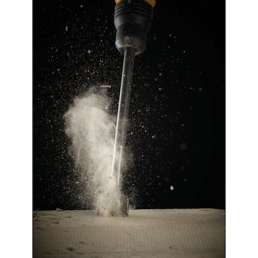 A close-up photo of a chisel drill bit striking a concrete surface, sending dust and debris into the air against a black background.