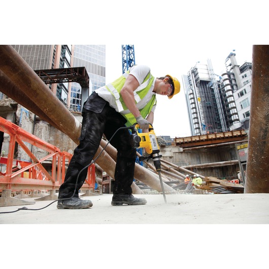 A person wearing a yellow hard hat and safety vest uses a DEWALT electric drill at a construction site with metal beams, concrete surfaces, and buildings in the background.