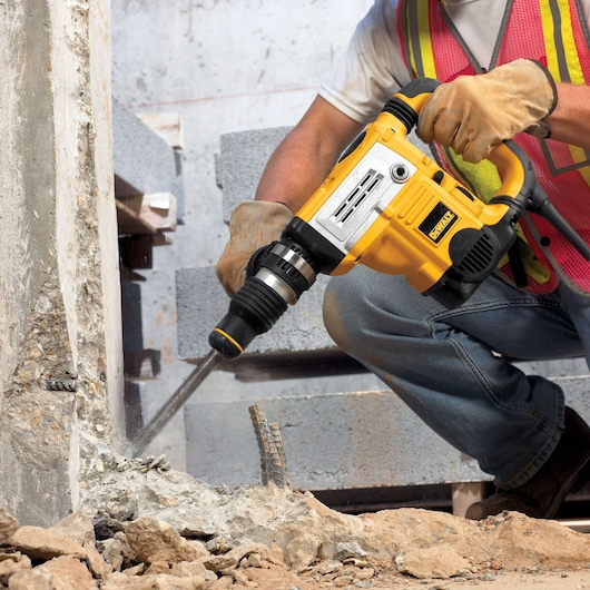 A person wearing work gloves and a safety vest is using a yellow DEWALT demolition hammer to break concrete at a construction site.