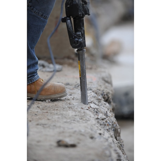 Close-up of a person using a power demolition tool with a chisel bit on a concrete surface. The person is wearing jeans and tan work boots.