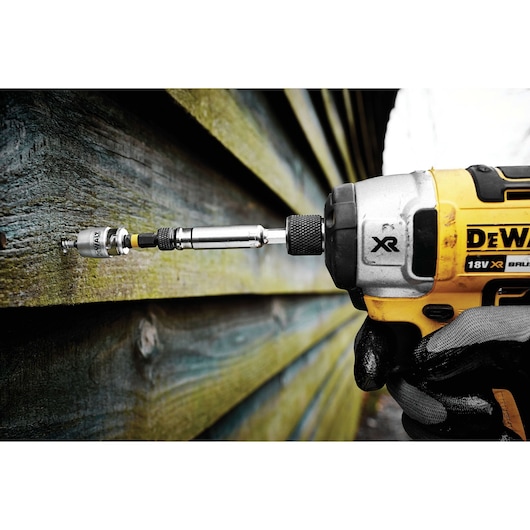 A close-up of a DEWALT cordless drill driving a screw into wooden planks, with a gloved hand holding the tool.