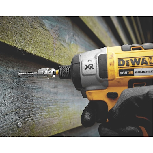 A close-up of a DEWALT 18V XR brushless power tool being used to drive a screw into a wooden panel. The tool is held by a gloved hand.