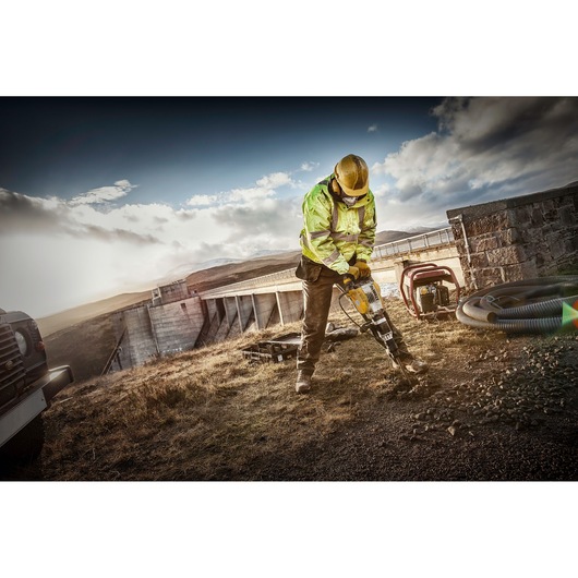 Worker uses DEWALT tool to break ground outdoors, wearing safety gear near a dam and construction equipment.
