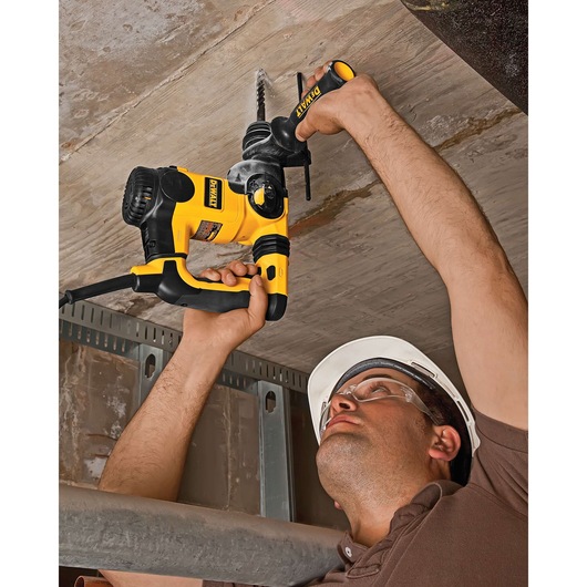 A person wearing a hard hat is using a yellow and black DEWALT power drill to drill into a concrete ceiling. The drill is plugged in, and the person's face is obscured.