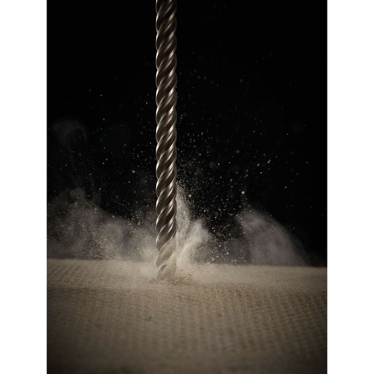 Metal drill bit penetrating a dusty surface, creating debris and dust against a dark background.