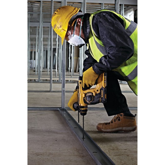 A construction worker wearing yellow protective gear, including a hard hat, gloves, safety vest, and face mask, uses a yellow DEWALT power drill to drill into a metal frame on a construction site.
