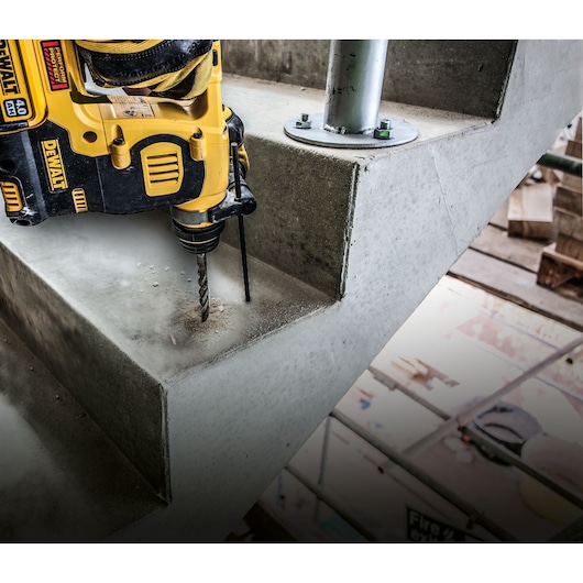 A DEWALT power drill is being used to drill into a concrete stair, creating dust. The drill is yellow and black and is positioned near a metal support post, illustrating construction or renovation work.