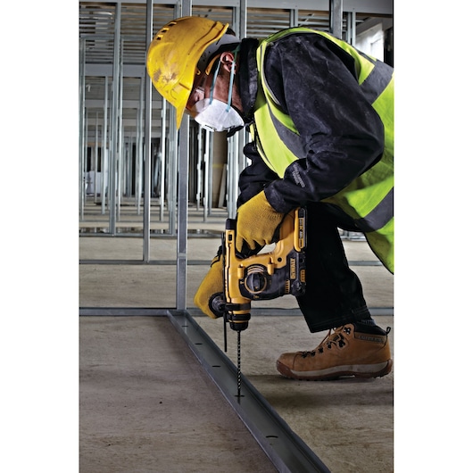 A construction worker wearing a yellow helmet, safety vest, gloves, and mask uses a DEWALT power drill to drill into a metal frame on a construction site.