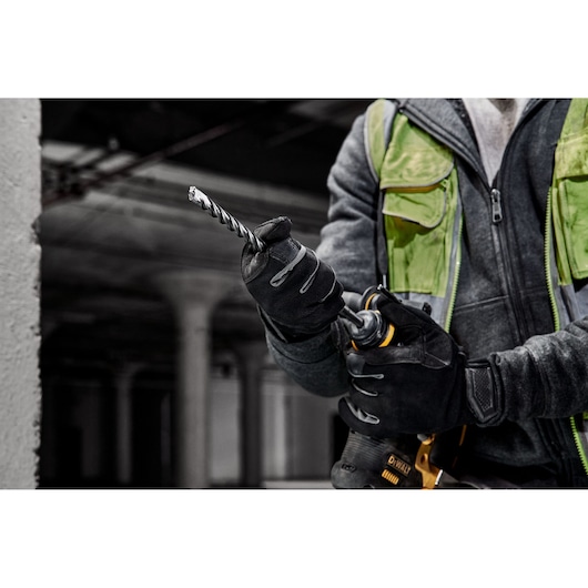 A person wearing gloves and a green safety vest is holding a DEWALT power drill with a spiral drill bit attached, in an industrial setting.