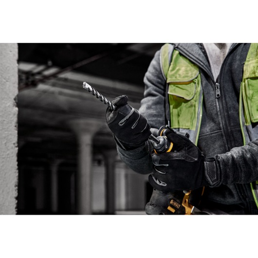 A person wearing work gloves and a green safety vest is holding a DEWALT power drill with a spiral drill bit, preparing for use at a construction site.