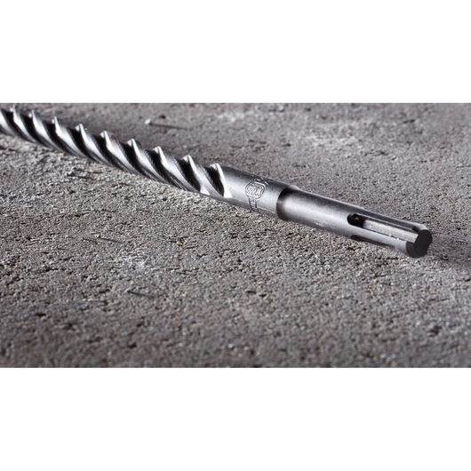 A close-up image of a DEWALT DT8972 drill bit with a spiral design and SDS shank, lying on a textured concrete surface.