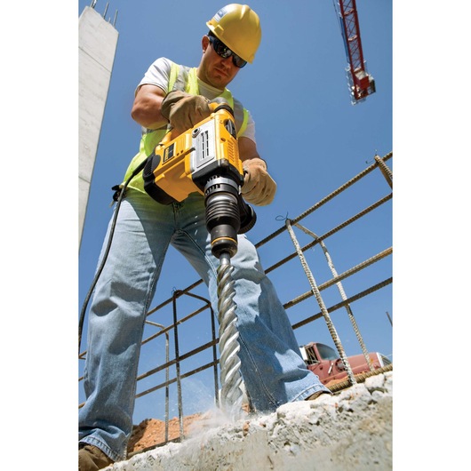 A construction worker, wearing a yellow safety helmet and gloves, uses a DEWALT power drill with an industrial drill bit to drill into concrete at a construction site. Steel reinforcement bars and construction equipment are visible in the background.