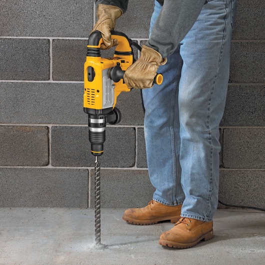 A person wearing gloves, jeans, and work boots uses a yellow DEWALT rotary hammer drill to drill into a concrete floor.