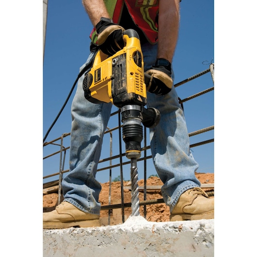 A person wearing work gloves and boots uses a yellow DEWALT rotary hammer drill to drill into concrete at a construction site. The drill bit is making a hole in the concrete, with rebar visible in the background.