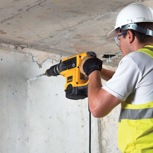 A person wearing safety gear is using a DEWALT electric drill with a long drill bit to bore into a concrete wall.