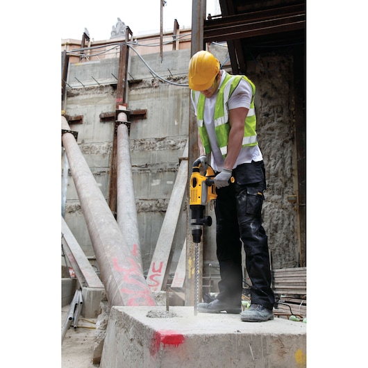 Worker uses DEWALT hammer drill DT9445 on concrete at construction site, wearing safety gear.